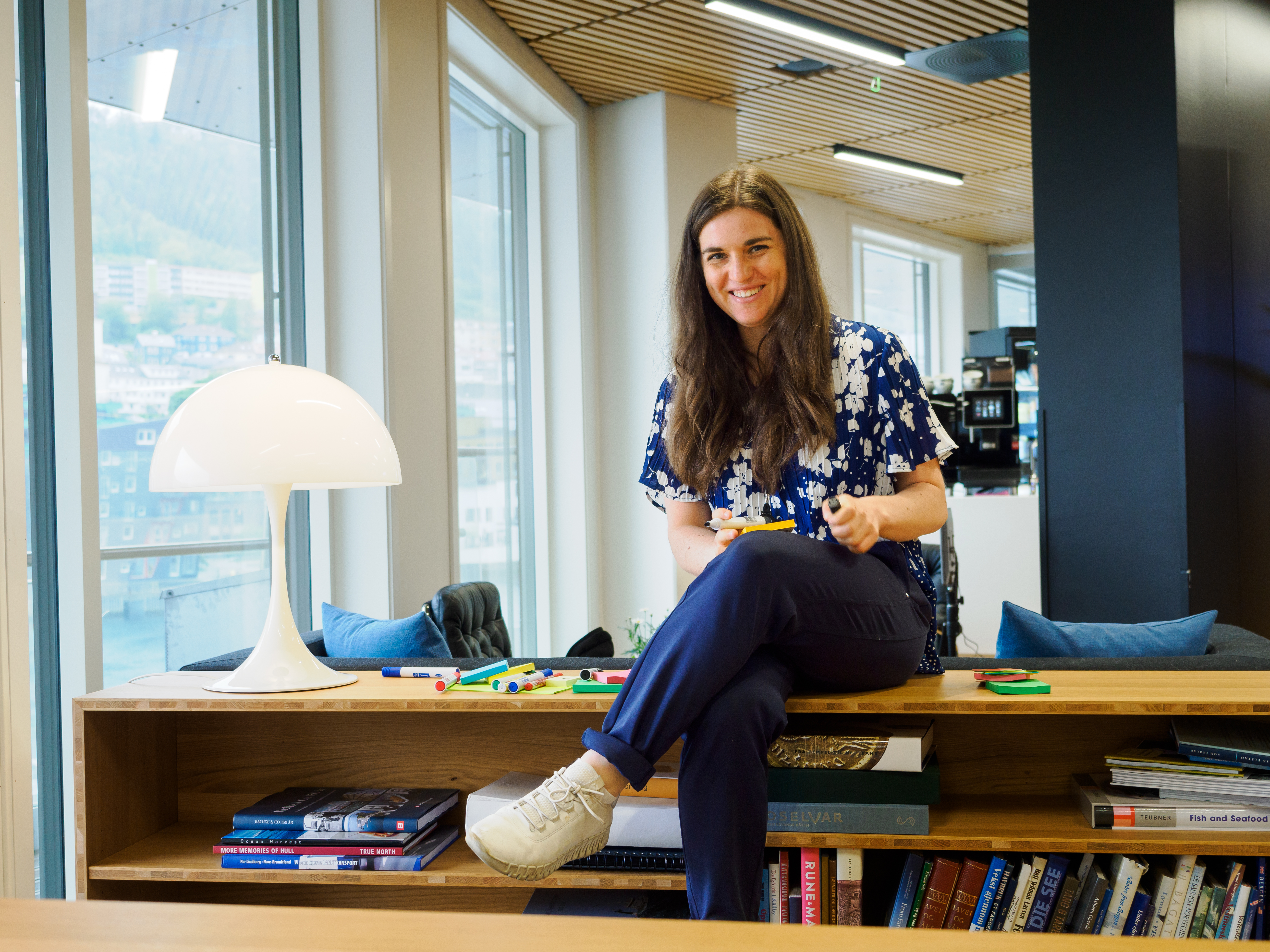 A brown haired woman smiling as she sits on top of a low book shelf.